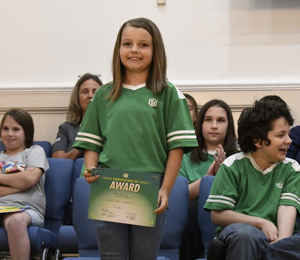 Young girl in a green shirt stands holding a certificate, with other children seated in the background, during an indoor event or ceremony.