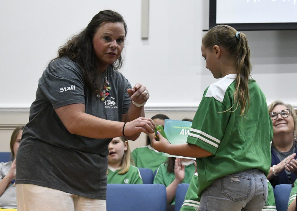 A woman hands an award or object to a girl in a green shirt during an indoor event, with others seated in the background.