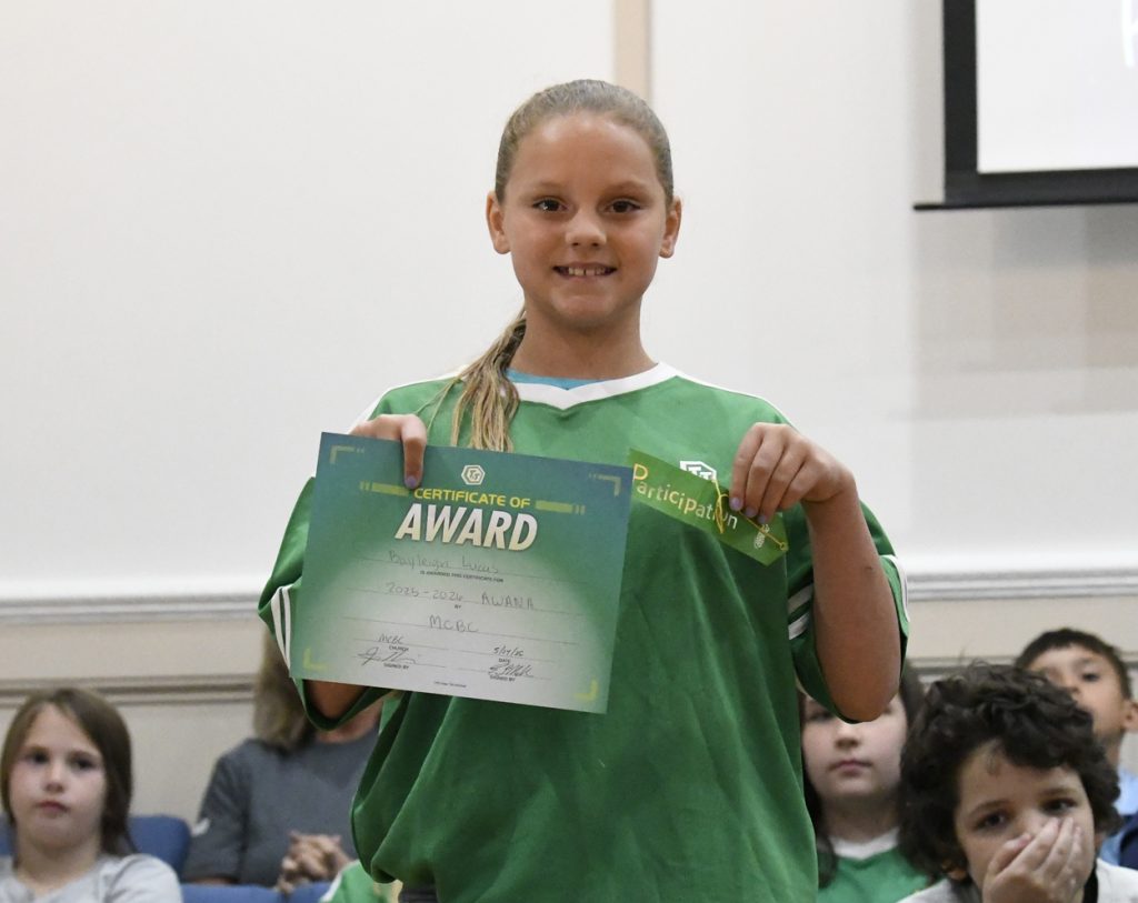 Girl in green shirt smiling and holding an award certificate in a classroom or assembly setting, with other students seated in the background.
