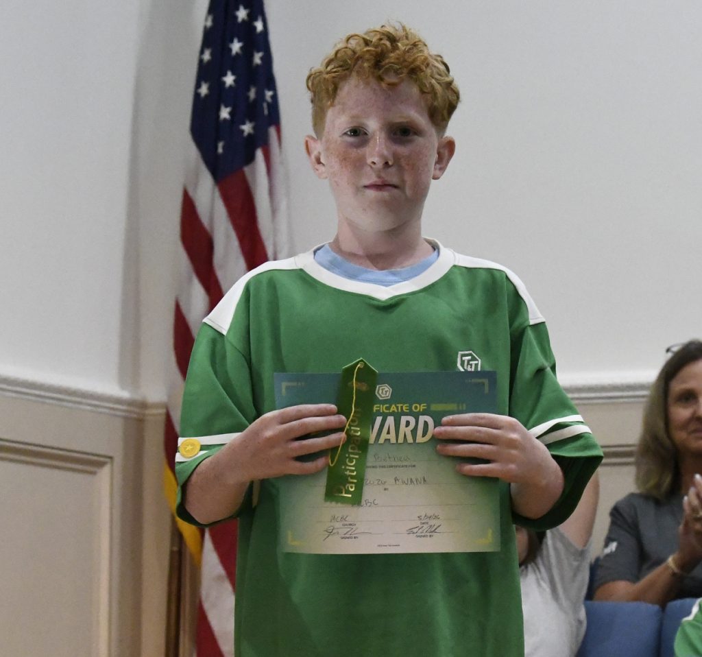 A boy with red hair in a green shirt holds an award certificate and ribbon, standing indoors in front of an American flag and a seated person.