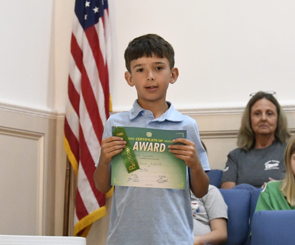 Young boy holding a certificate and ribbon, standing in front of a U.S. flag, with people seated in the background.