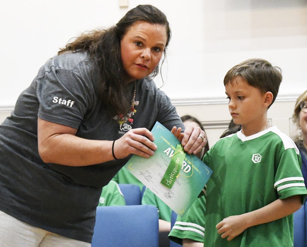 Woman presenting a certificate to a young boy wearing a green shirt, both standing indoors with other children in the background.