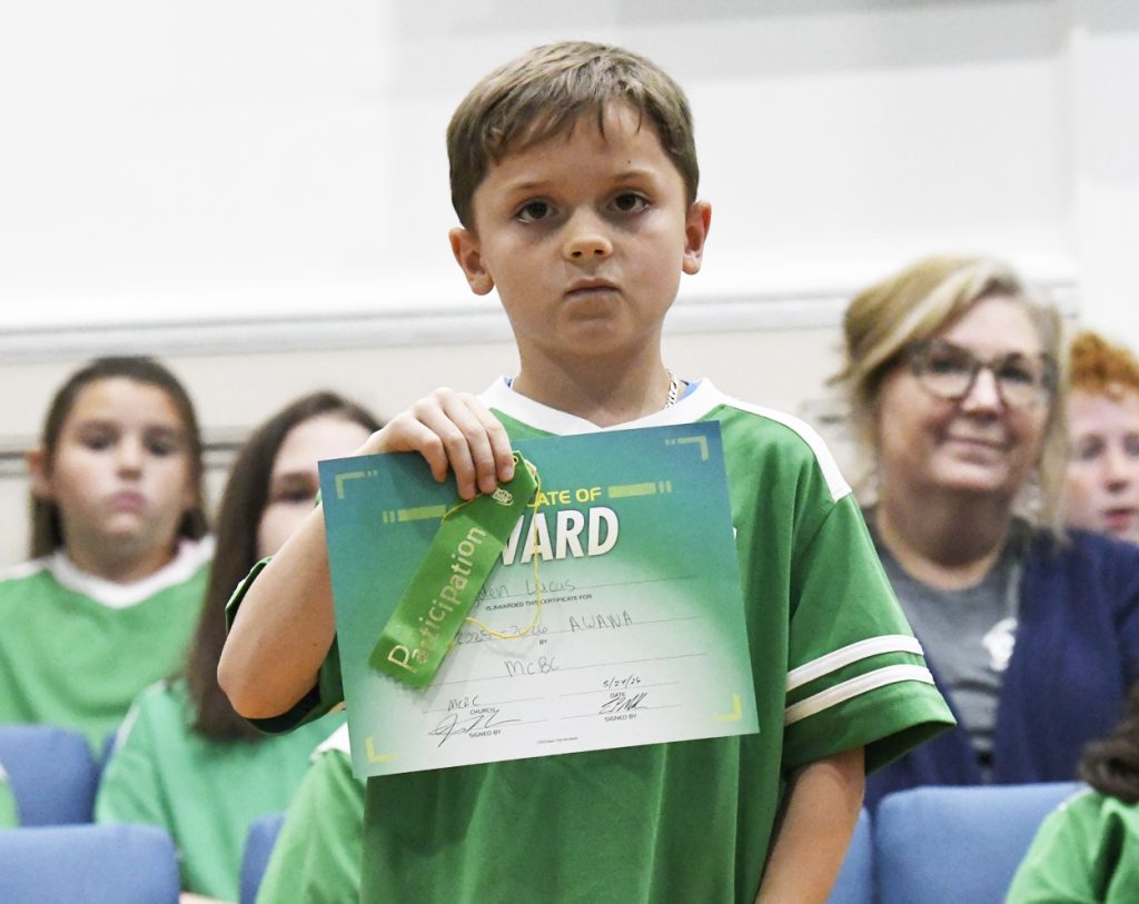 A young boy in a green shirt holds up an award certificate and ribbon, standing indoors with several seated people in the background.