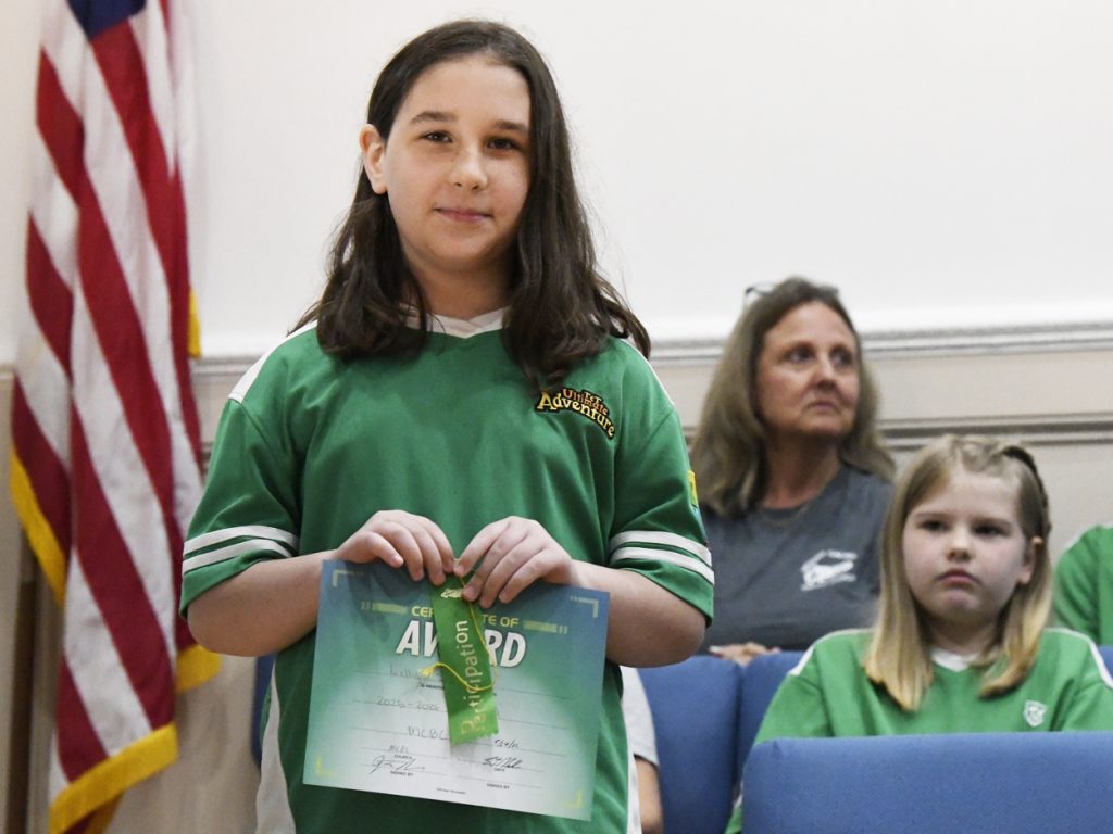 A child in a green shirt holds an award certificate and ribbon, standing indoors with an American flag in the background and people seated nearby.