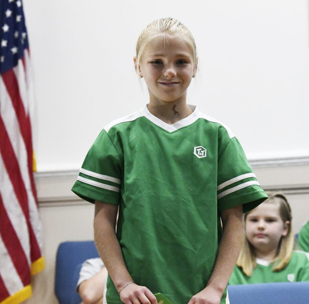 A young girl in a green shirt stands in front of an American flag, with other children seated in the background.