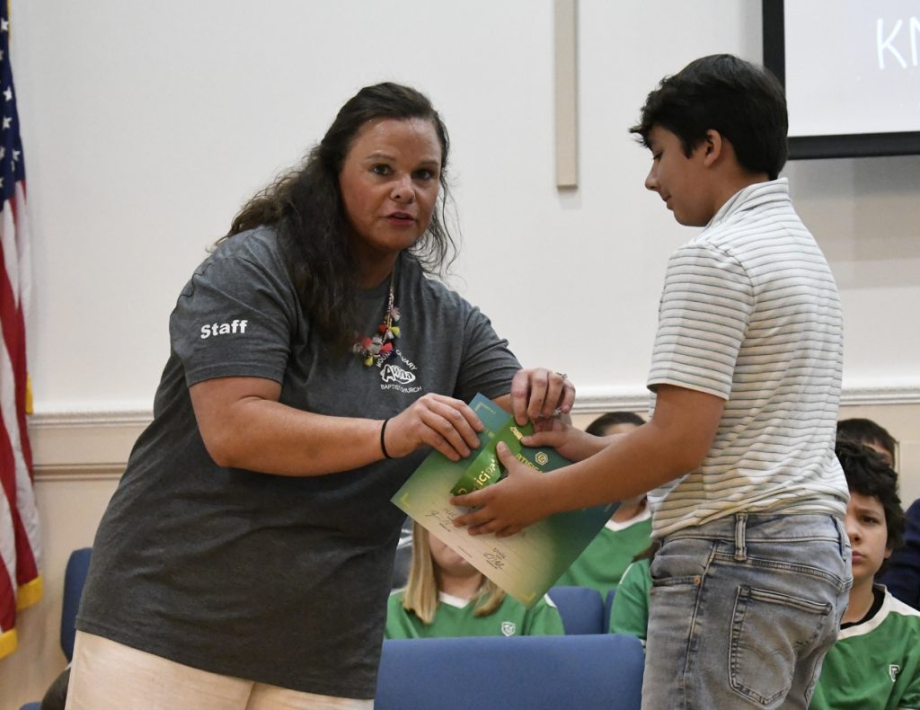 A woman hands a certificate or folder to a student during an indoor event or ceremony, with people seated in the background.