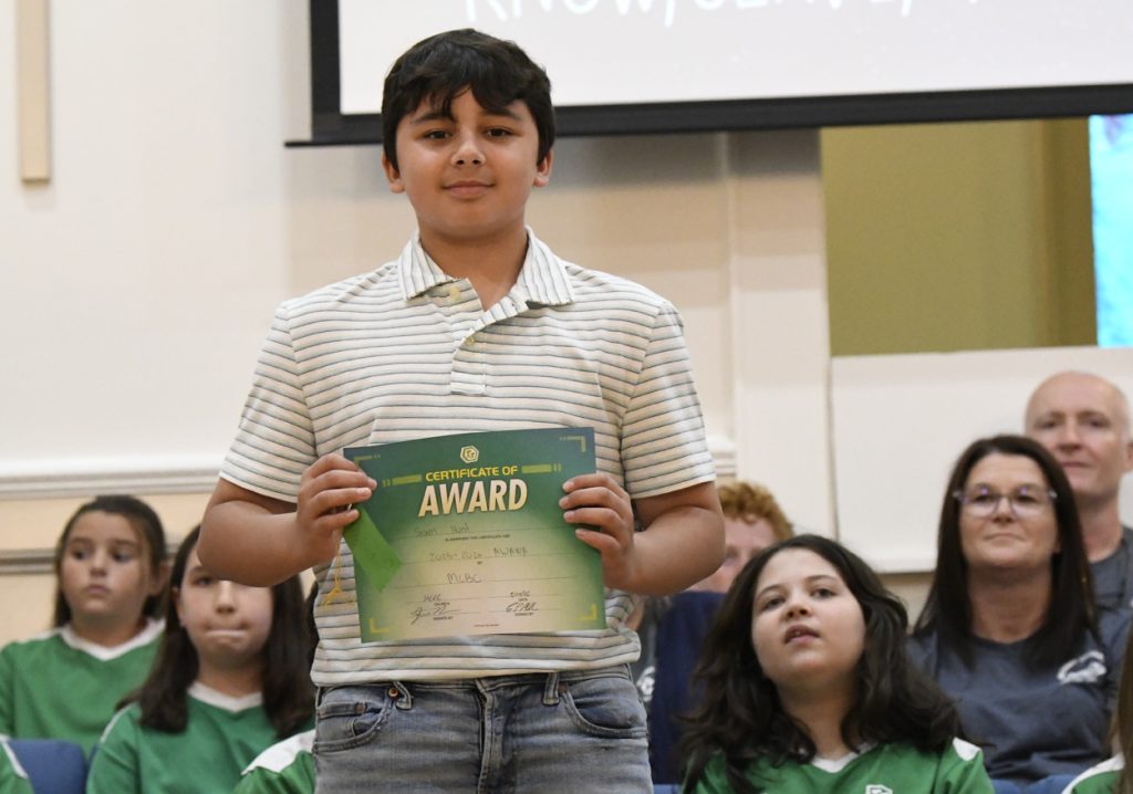 A boy stands holding an award certificate, with other children seated in the background on a stage.