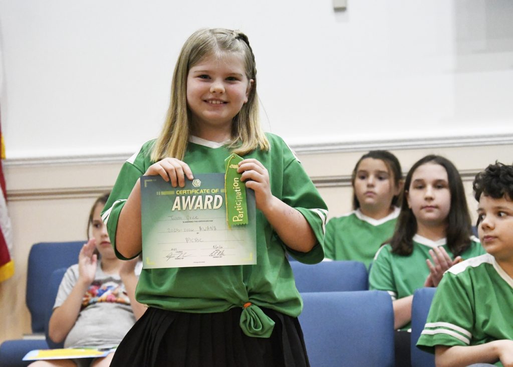 Smiling girl in green shirt holds an award certificate and ribbon, standing in front of seated children who are clapping.