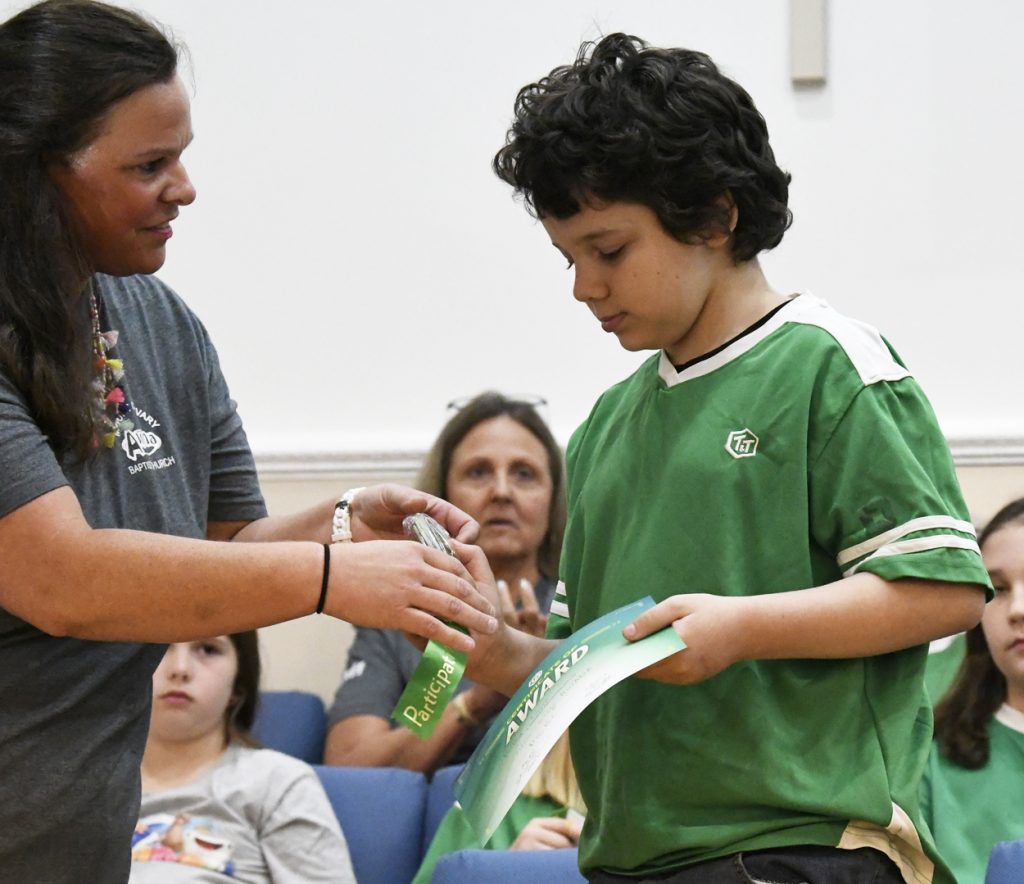 A woman hands an award to a boy in a green shirt during an indoor ceremony, while people watch in the background.