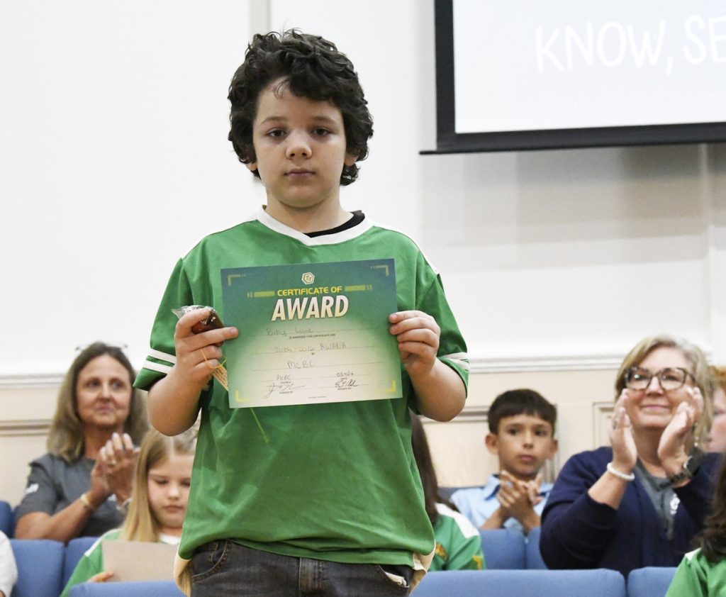 A young boy holding an award certificate stands in front of a seated audience, some of whom are clapping in support.