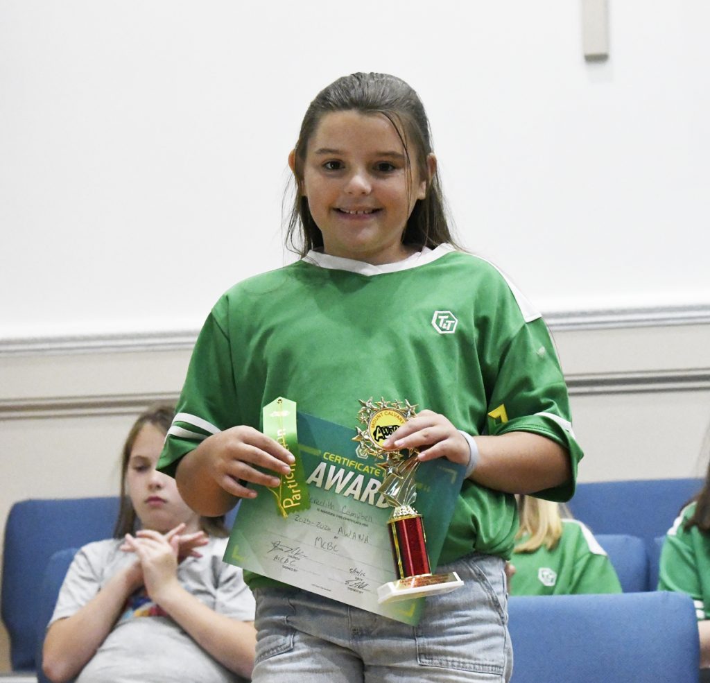 Girl in green shirt holding an award certificate and trophy, smiling, standing indoors with other children seated in the background.