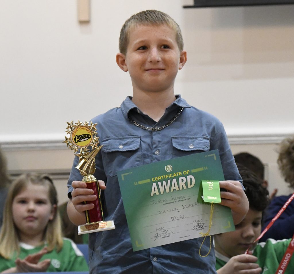 A boy holding a trophy and an award certificate, smiling at a ceremony, with other children seated in the background.