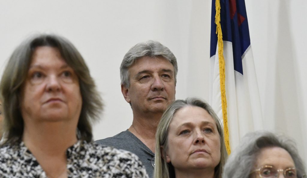 Three adults standing indoors, two women in the foreground and a man in the background, with a flag visible behind them.