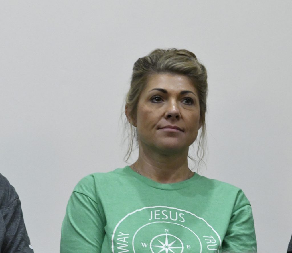 Woman with blonde hair wearing a green shirt with "JESUS" printed on it, standing against a plain light-colored background.