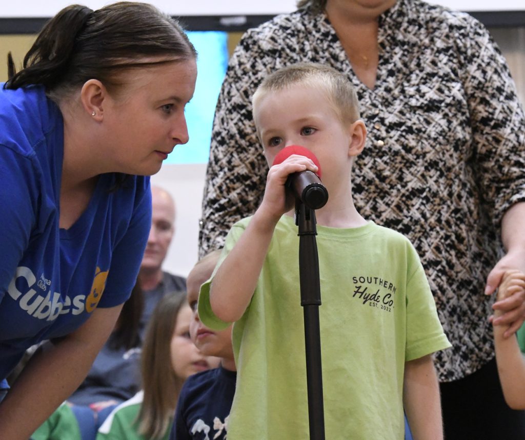 A young boy speaks into a microphone while a woman crouches beside him, listening. Other people are visible in the background.