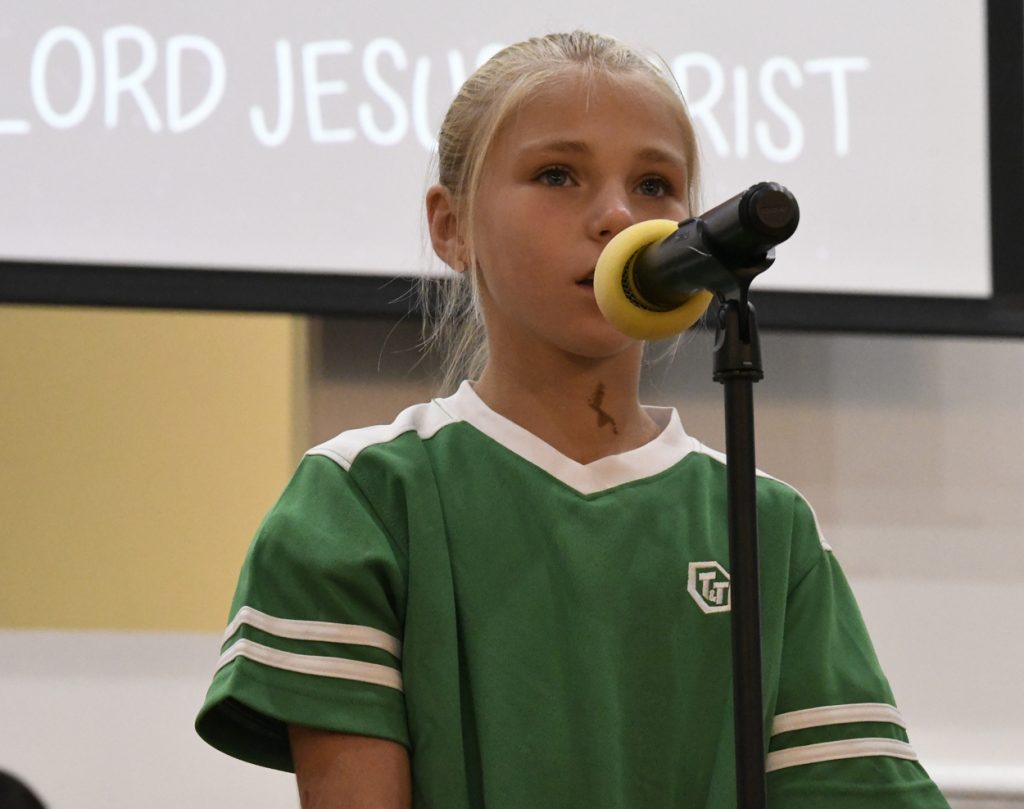 A young girl in a green shirt speaks into a microphone on stage, with text about Jesus Christ visible on the screen in the background.