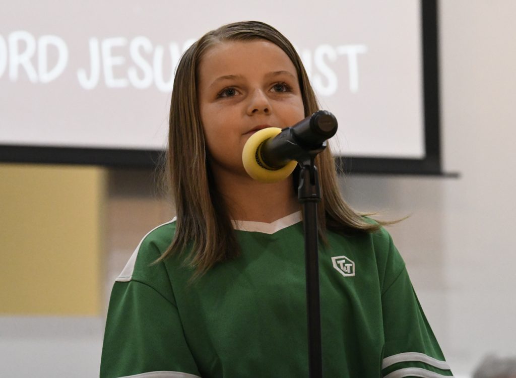 A young girl with straight brown hair speaks into a microphone, wearing a green shirt, with "JESUS CHRIST" partially visible on a screen behind her.