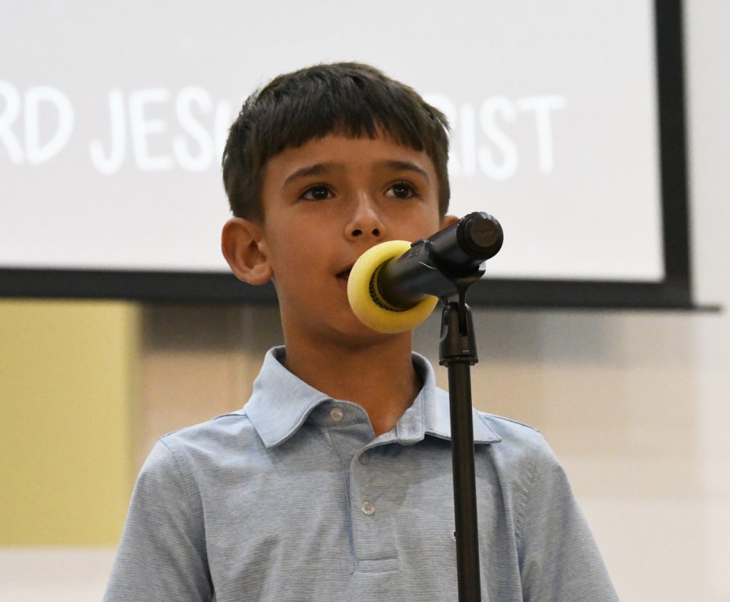 A young boy in a light blue shirt speaks into a microphone with a yellow windscreen, standing indoors with a blurred screen in the background.