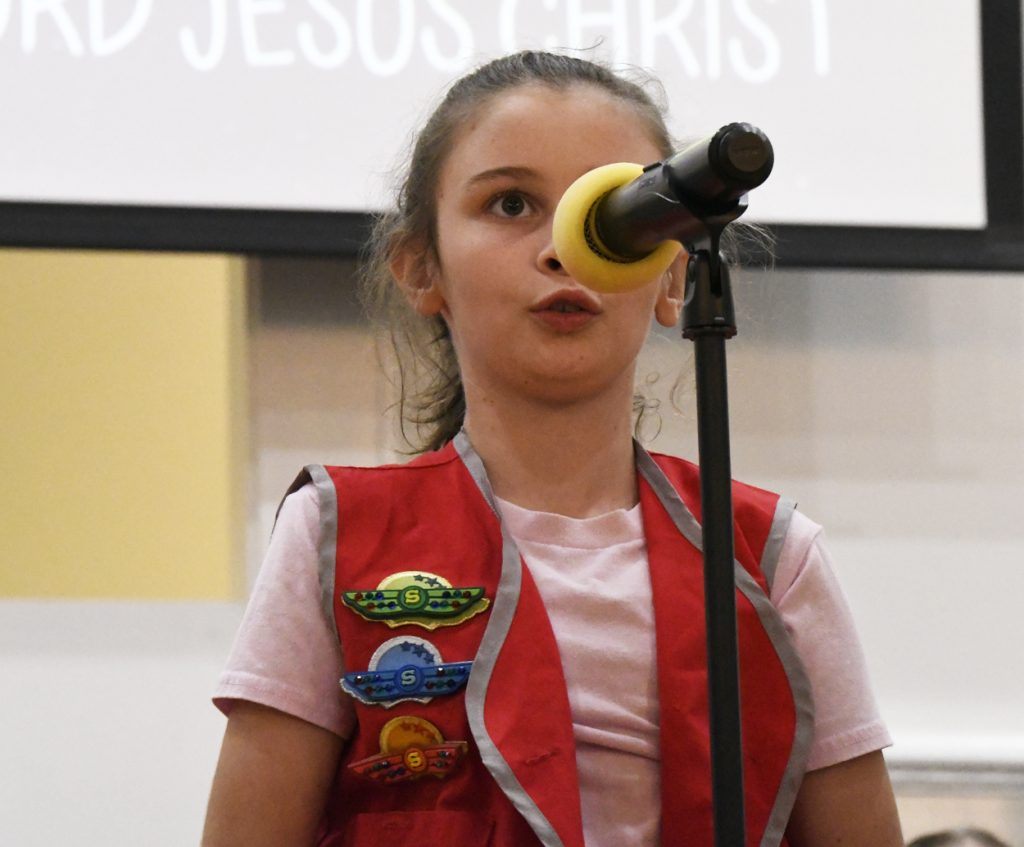 Young girl in red vest with badges speaks into a microphone on a stage, a screen with partial text visible in the background.