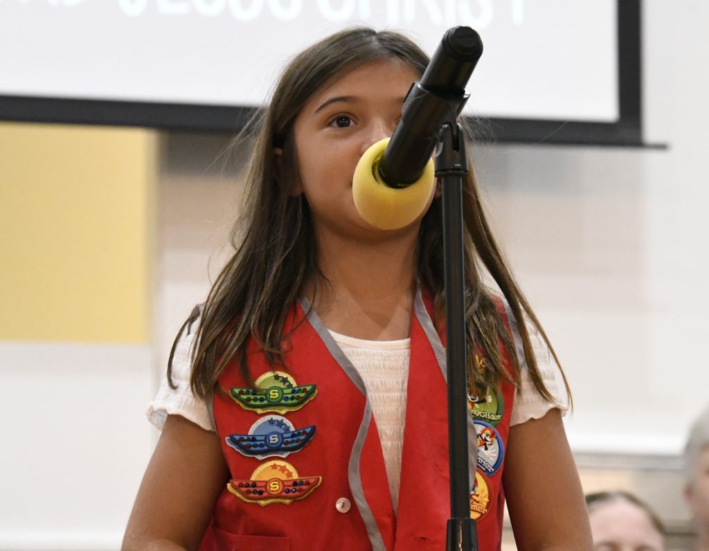 A young girl wearing a red vest with colorful badges speaks into a microphone at an indoor event.