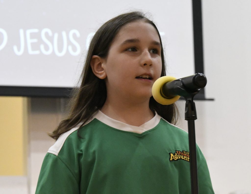 A young person with long hair speaks into a microphone on a stand, wearing a green shirt, with a blurred screen in the background.