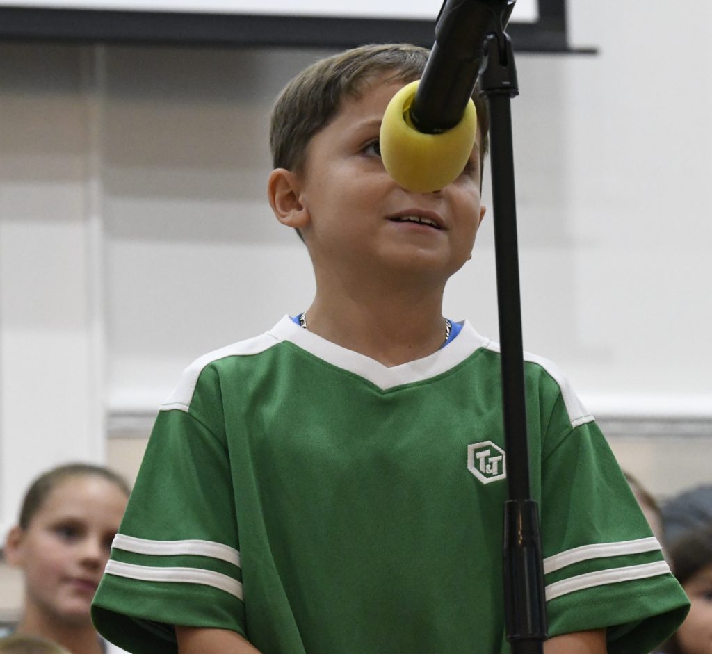 Young boy in a green shirt speaking into a microphone with a yellow cover, with blurred people and a screen in the background.