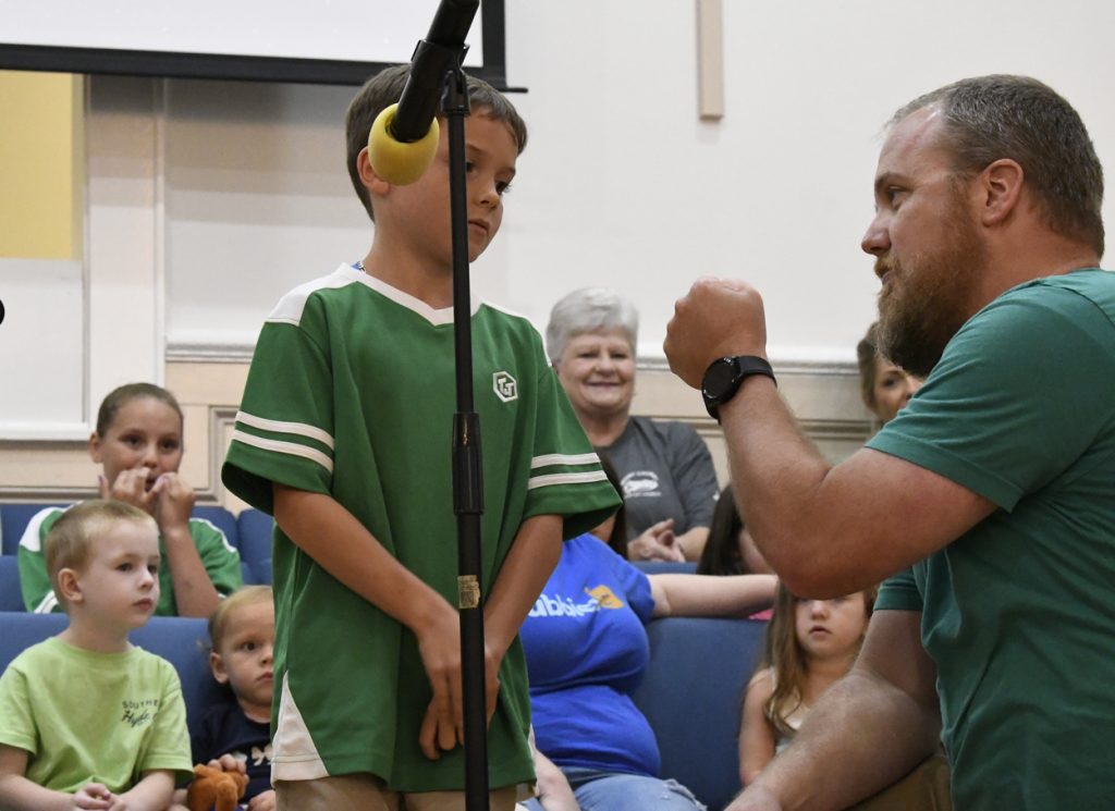 A boy in a green shirt stands by a microphone while a man kneels and gestures, with several people seated and watching in the background.