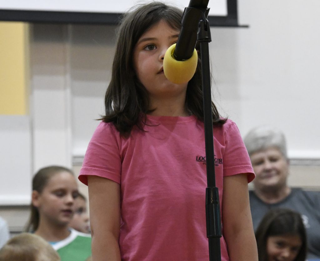 A young girl in a pink shirt stands in front of a microphone, with other children and adults visible in the background.