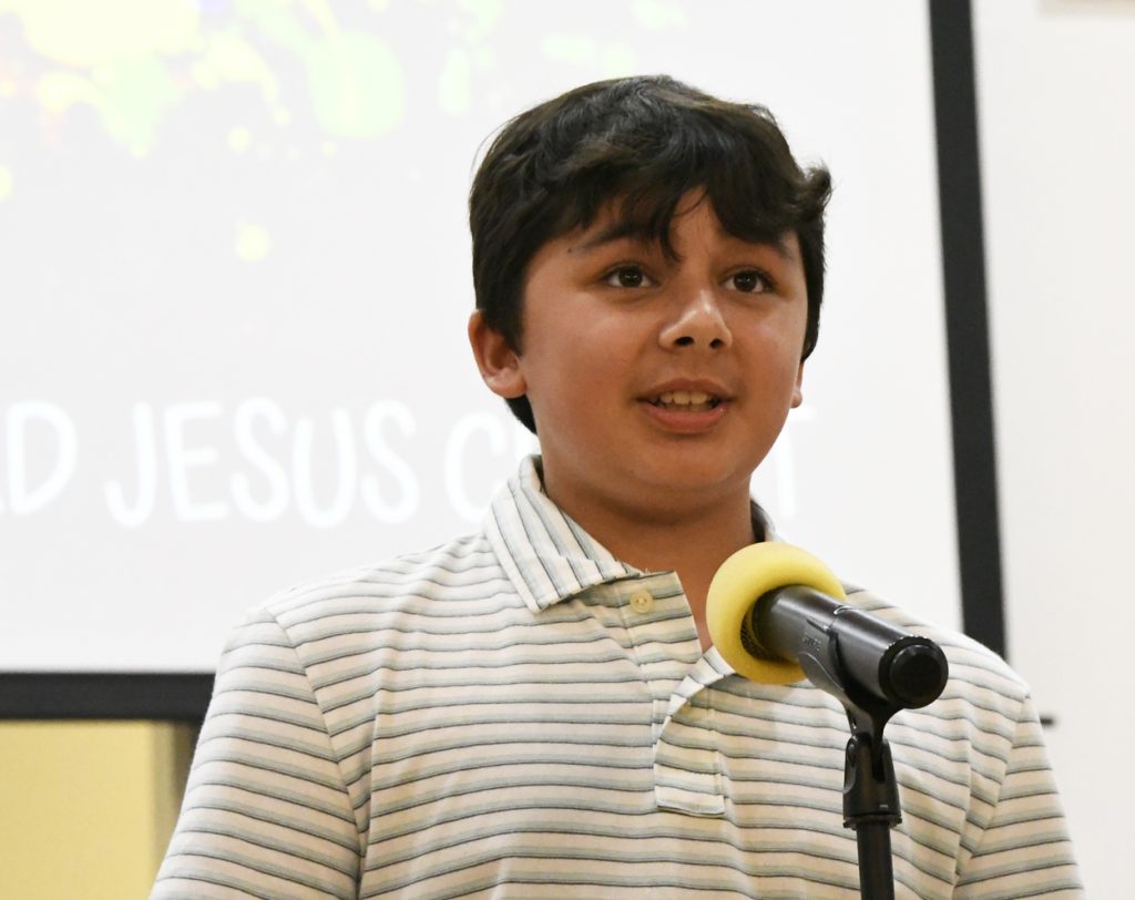 A boy in a striped shirt speaks into a microphone, standing in front of a screen displaying partially visible text.