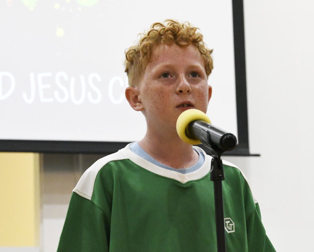 A young boy with curly red hair speaks into a microphone, wearing a green shirt, standing indoors with a screen in the background.