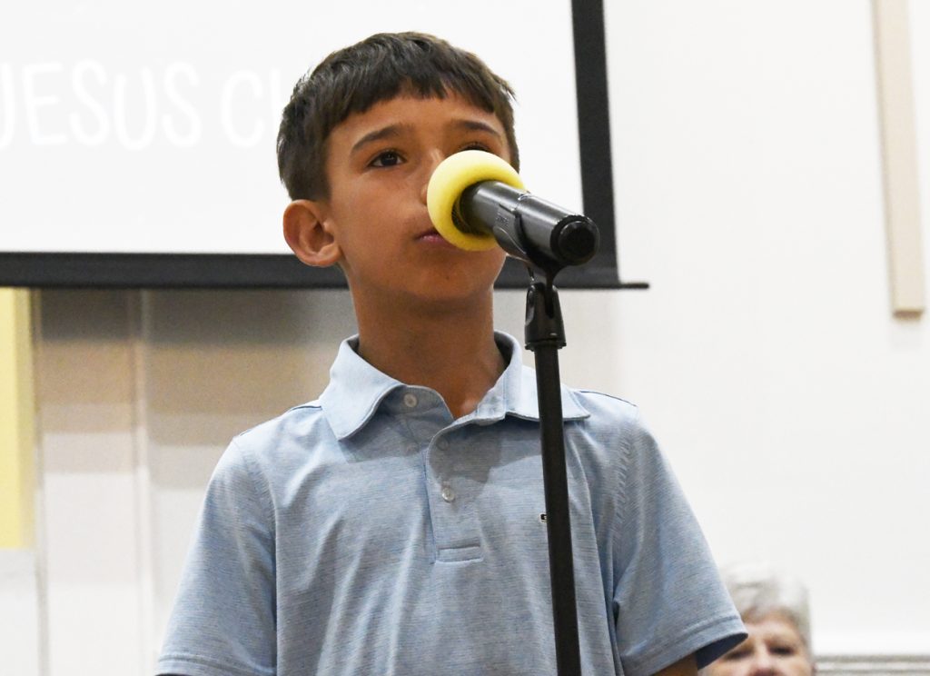 A young boy in a blue polo shirt speaks into a microphone, possibly participating in a spelling bee or presentation indoors.