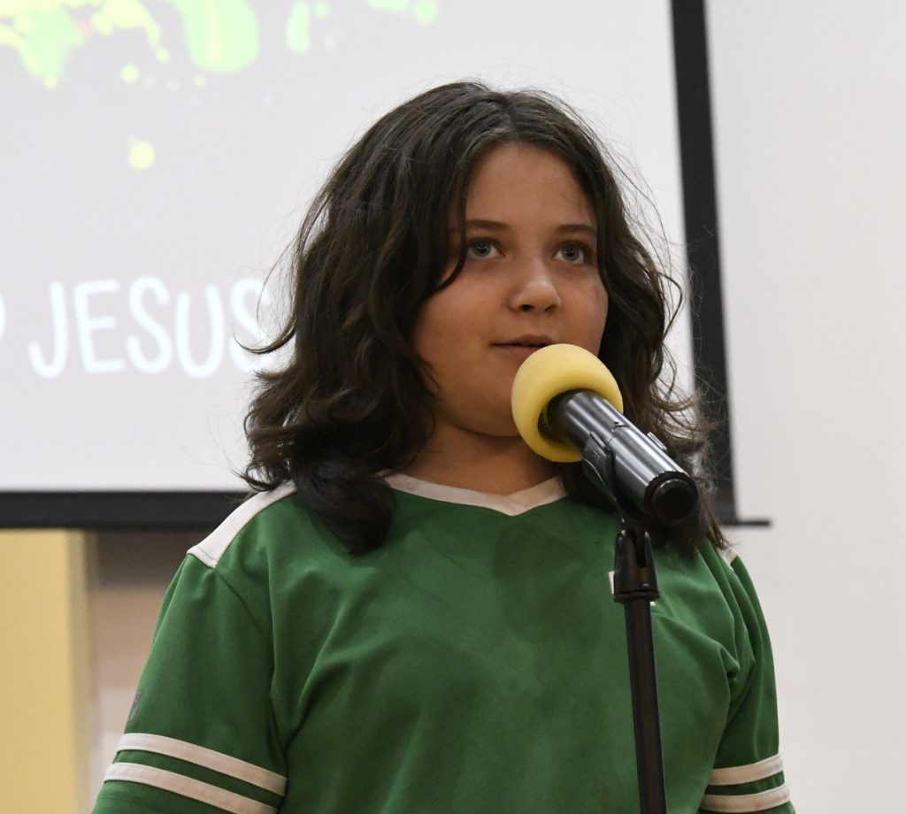 A child with long hair speaks into a microphone while standing indoors, wearing a green shirt and looking forward.