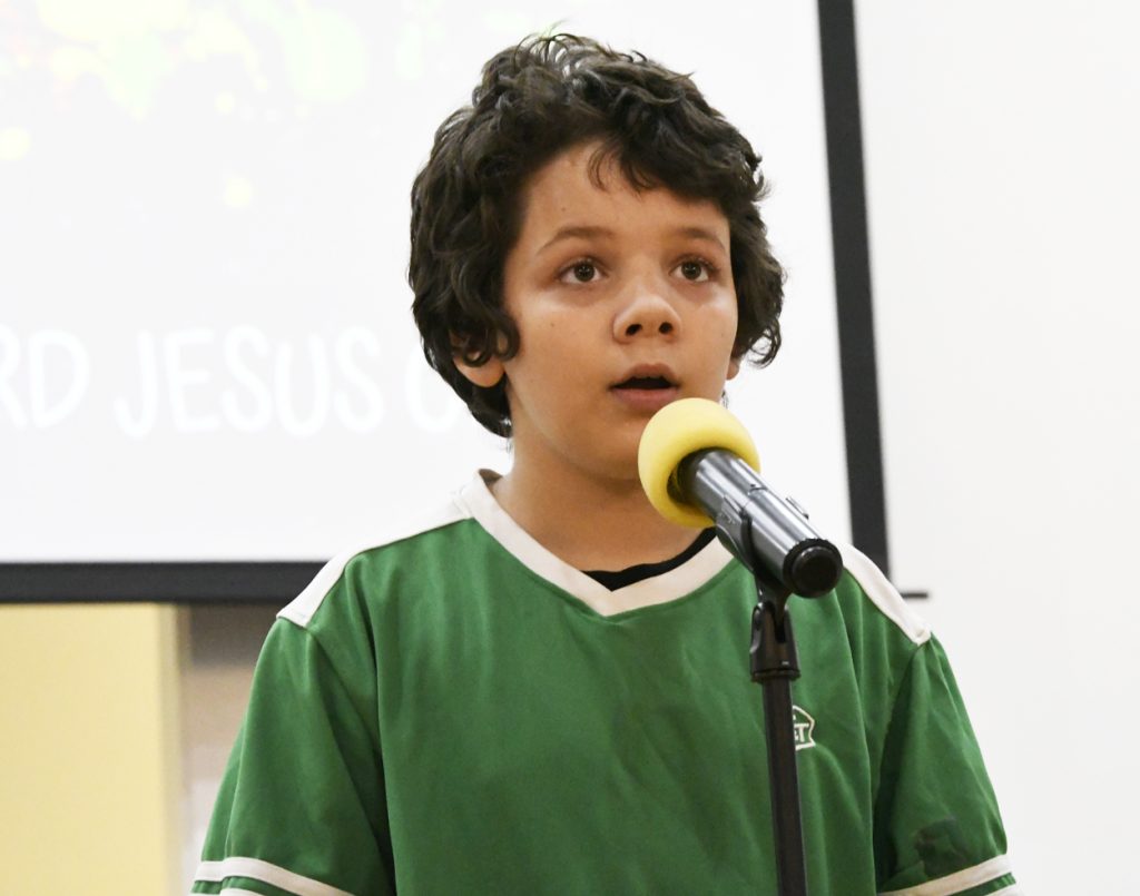 A young boy with curly hair wearing a green shirt speaks into a microphone on a stand, with a projected screen in the background.