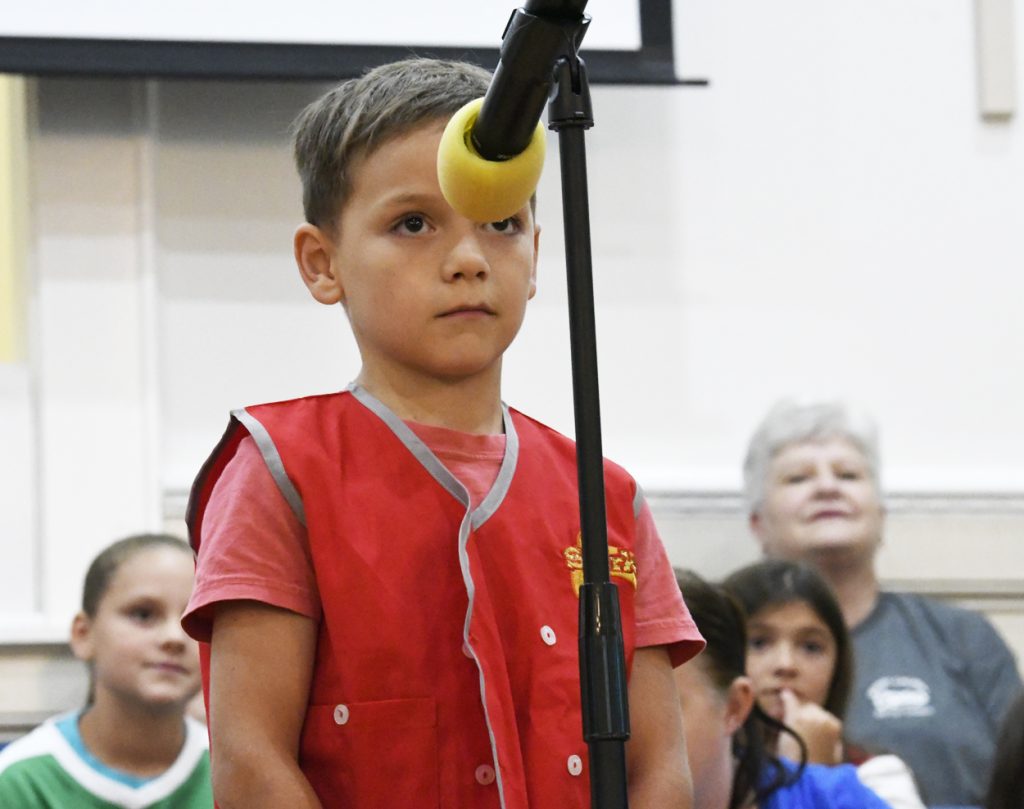 A young boy in a red vest stands at a microphone, with several people seated and watching in the background.