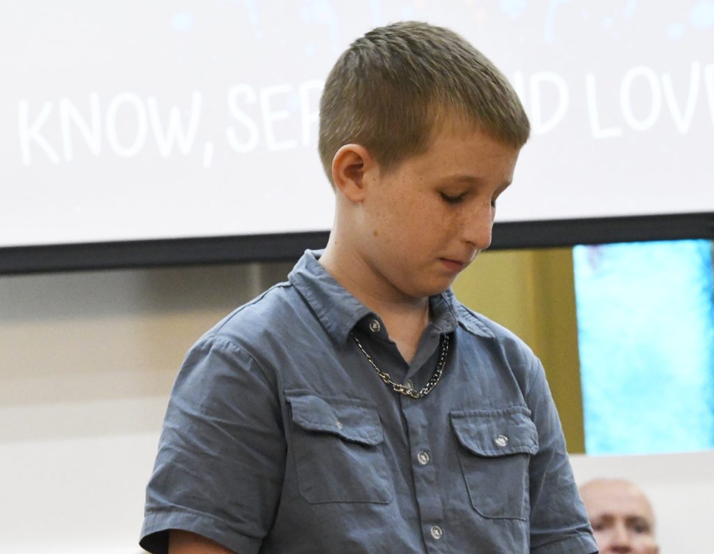 A young boy with short hair stands indoors, looking down, wearing a blue button-up shirt. A blurred screen with text appears in the background.