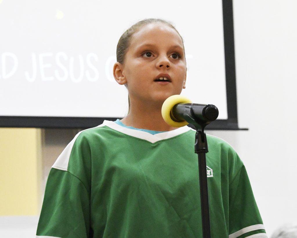 A young person in a green shirt speaks or sings into a microphone on a stand, with the word "JESUS" visible in the background.