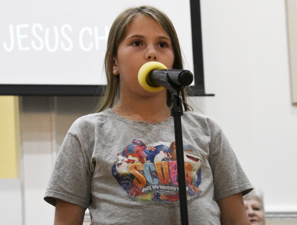A young girl with long hair stands at a microphone, speaking. She wears a graphic t-shirt and is indoors, possibly at an event or presentation.