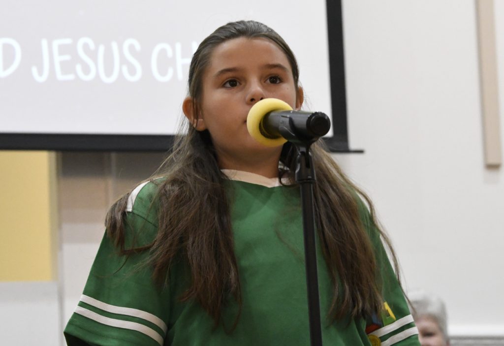 A young girl with long hair speaks or sings into a microphone, wearing a green shirt, standing indoors with a screen in the background.