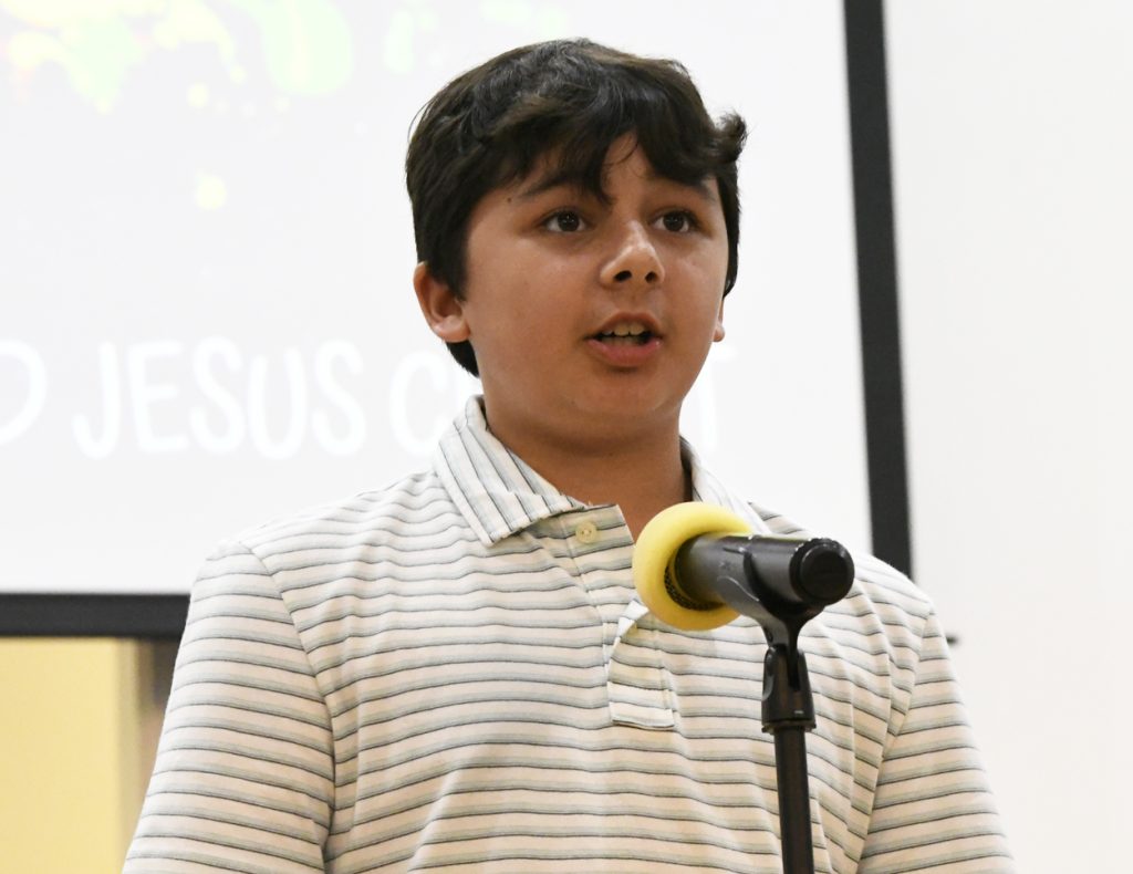 A boy wearing a white striped shirt speaks into a microphone on a stand, with a blurred presentation screen in the background.