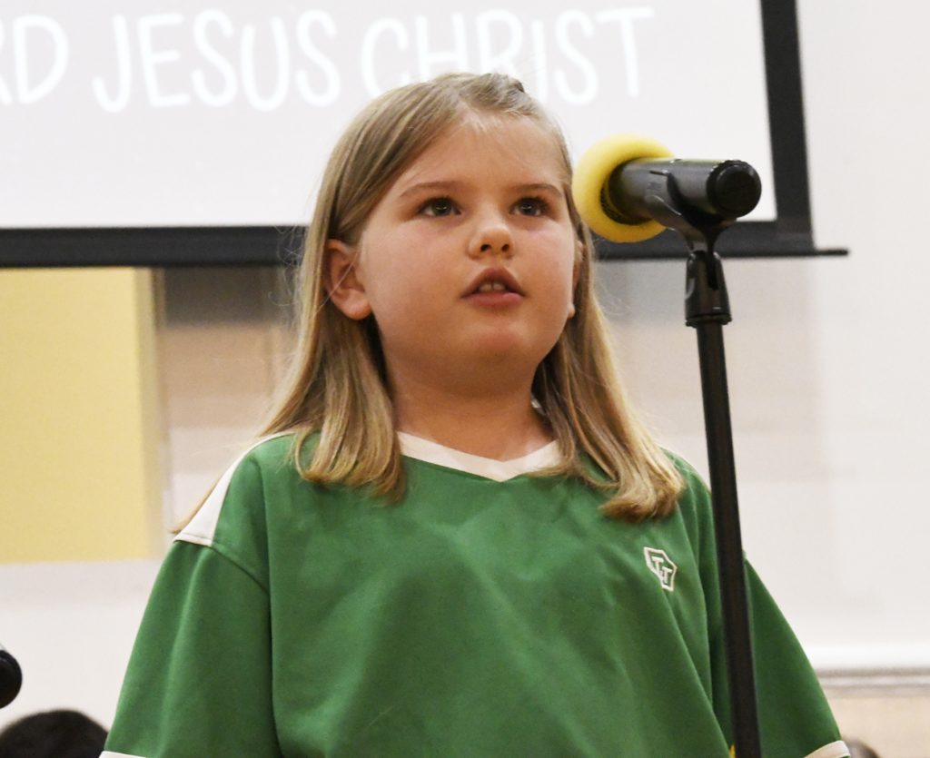 A young girl in a green shirt speaks or sings into a microphone, with a screen behind her displaying the words "JESUS CHRIST."