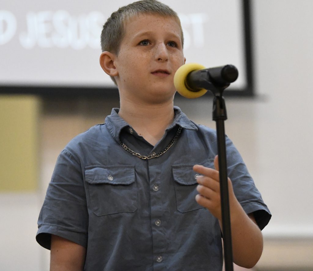 A young boy in a blue shirt speaks or sings into a microphone on a stand, standing indoors with a blurred projector screen behind him.