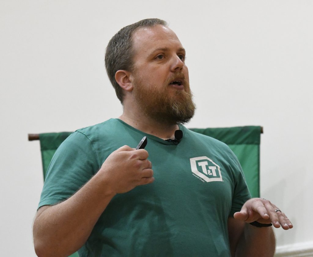 A man with a beard speaks while gesturing, wearing a green t-shirt and holding a marker or remote, standing indoors near a green banner.