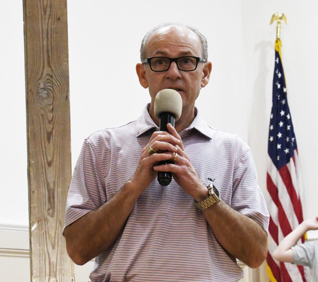 older man wearing glasses speaks into a microphone indoors, with a wooden post and a U.S. flag visible in the background.