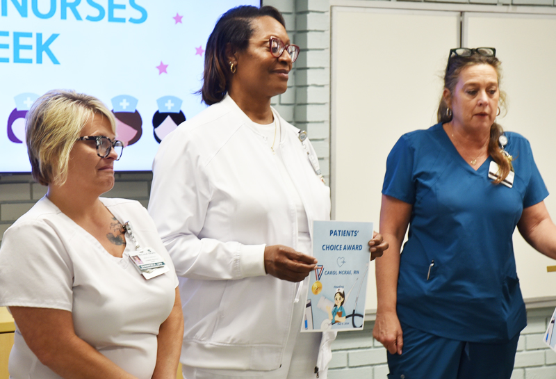 Three nurses standing indoors; one in the center holds a "Patients' Choice Award" certificate during a celebration for Nurses Week.