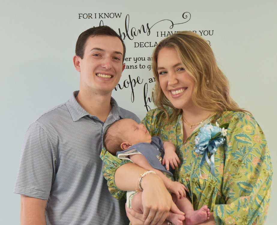 Smiling man and woman holding a sleeping baby, standing in front of a wall with inspirational black text.