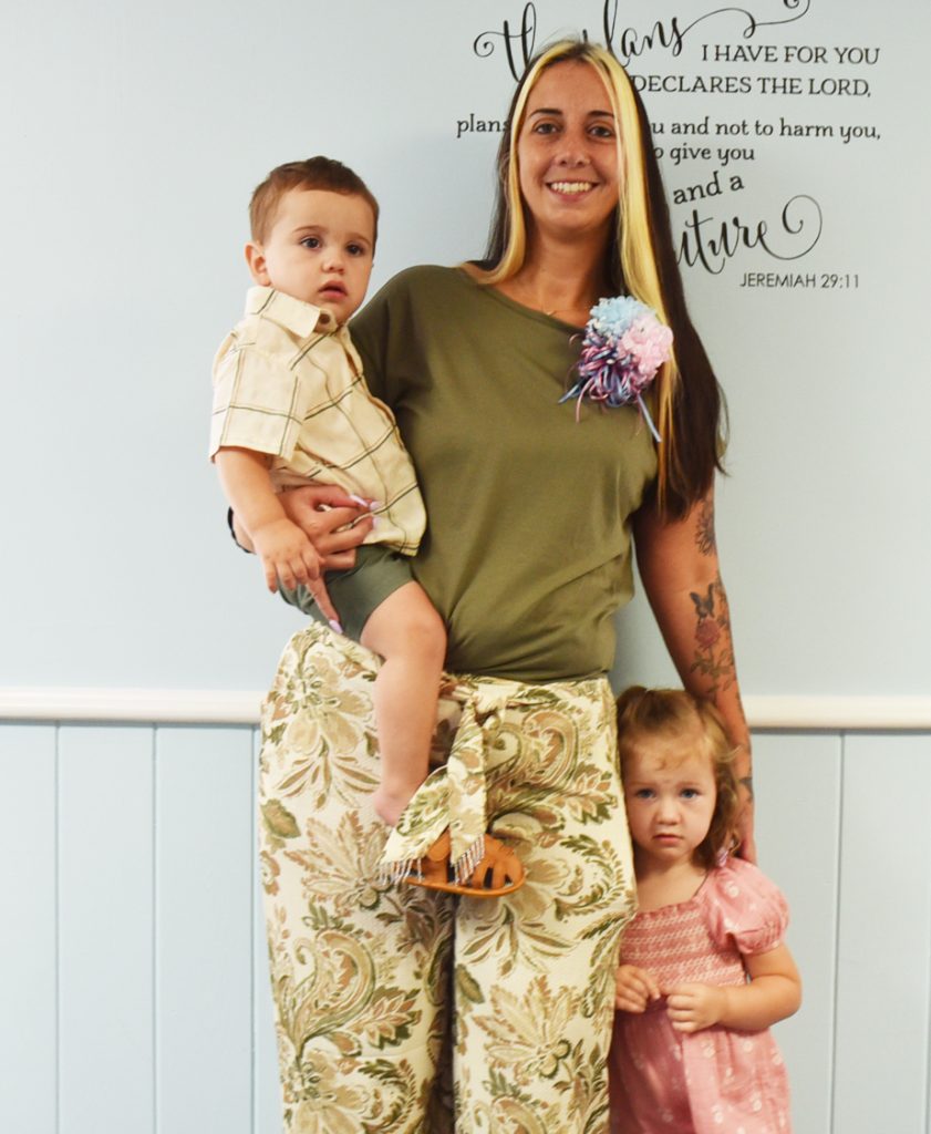 A woman stands smiling with a young boy in her arms and a young girl by her side, in front of a Bible verse wall decal.