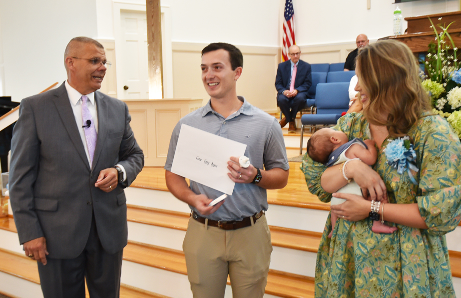 A man in a suit, a smiling man holding an envelope, and a woman holding a baby stand together in a church with people seated in the background.