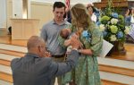 A man kneels and blesses a baby held by a woman, with another man standing beside them in a church setting.