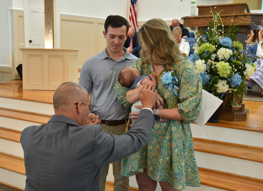 A man kneels and blesses a baby held by a woman, with another man standing beside them in a church setting.