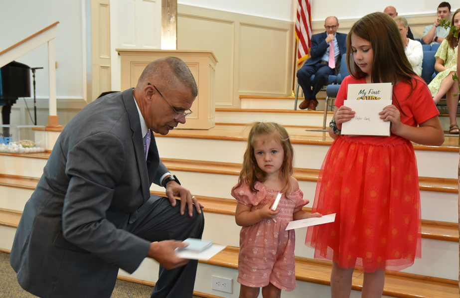 A man in a suit kneels, handing envelopes to two young girls on church steps; the older girl holds a book, and people watch in the background.